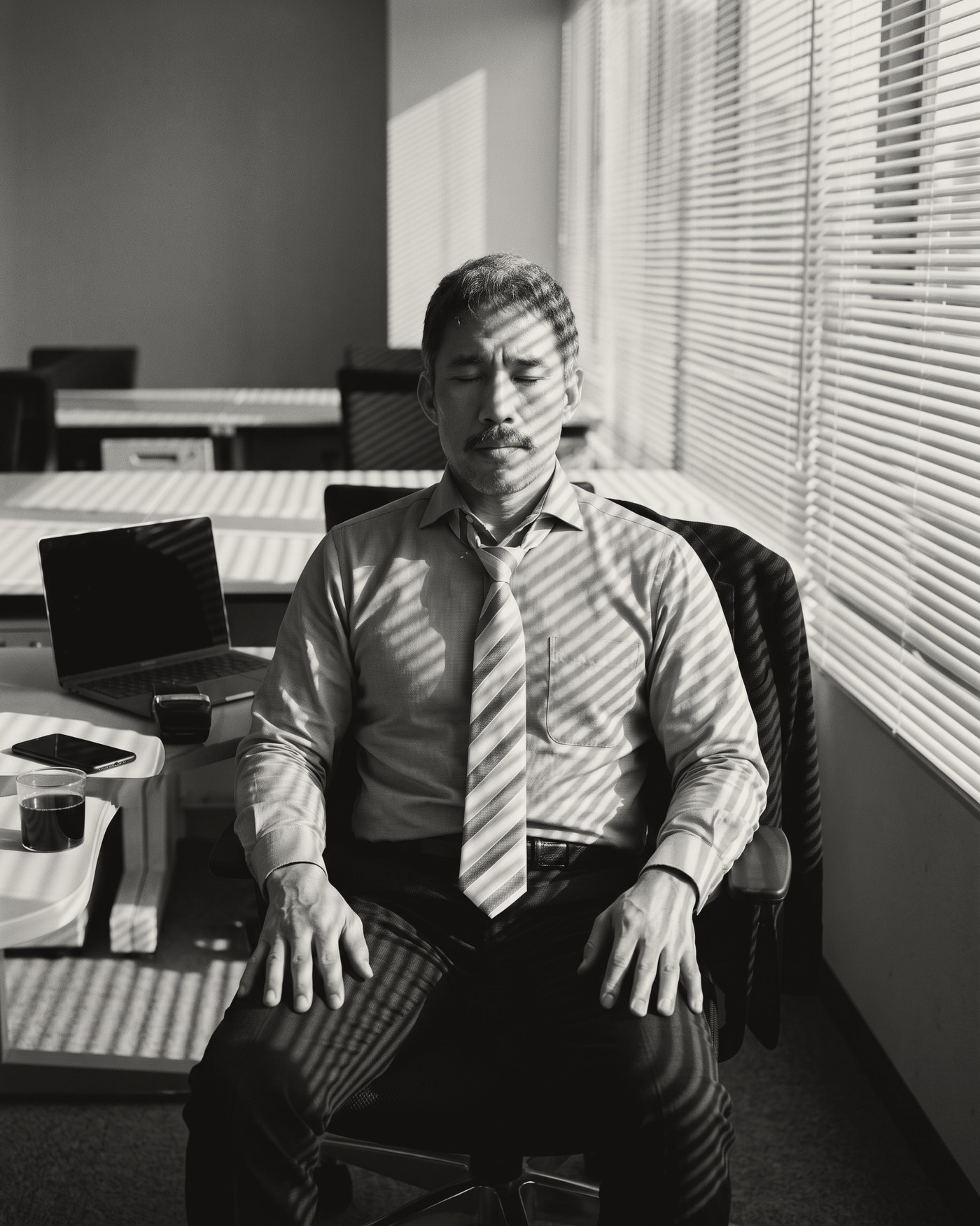 A man in shirt and tie sitting upright in his office chair with his eyes closed and hands on his thighs, light through the blinds striping him and the desk.