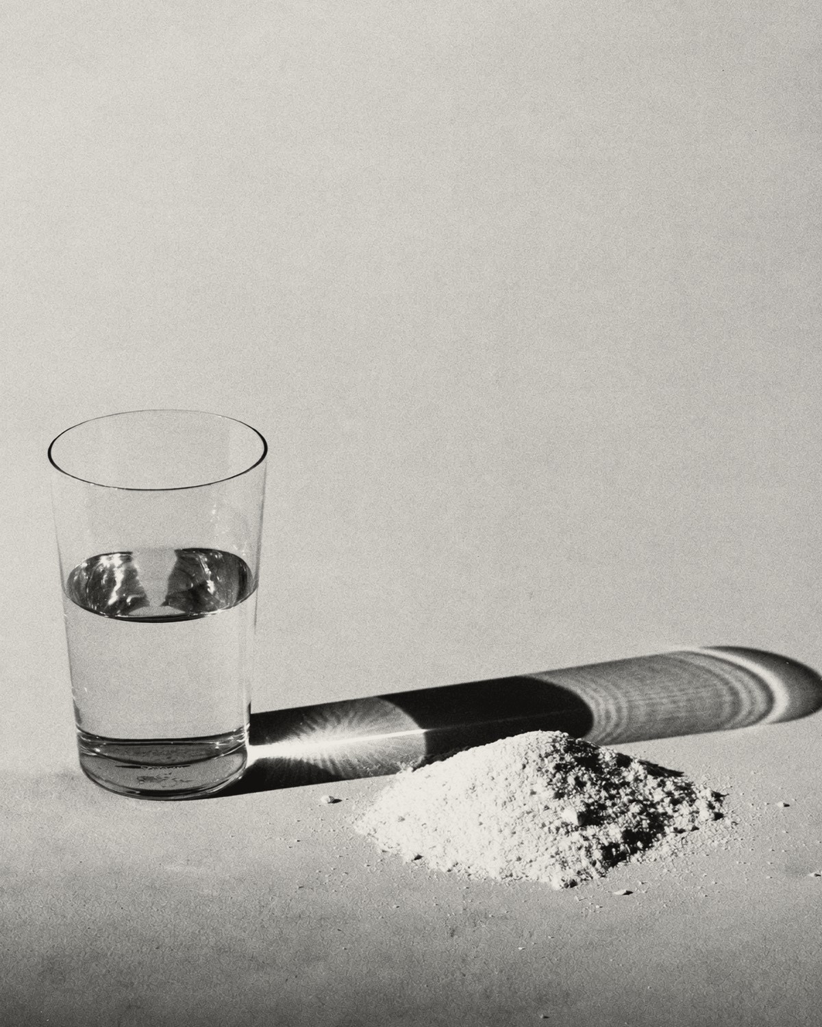A half-full glass of water casting a long shadow across a small heap of white powder.