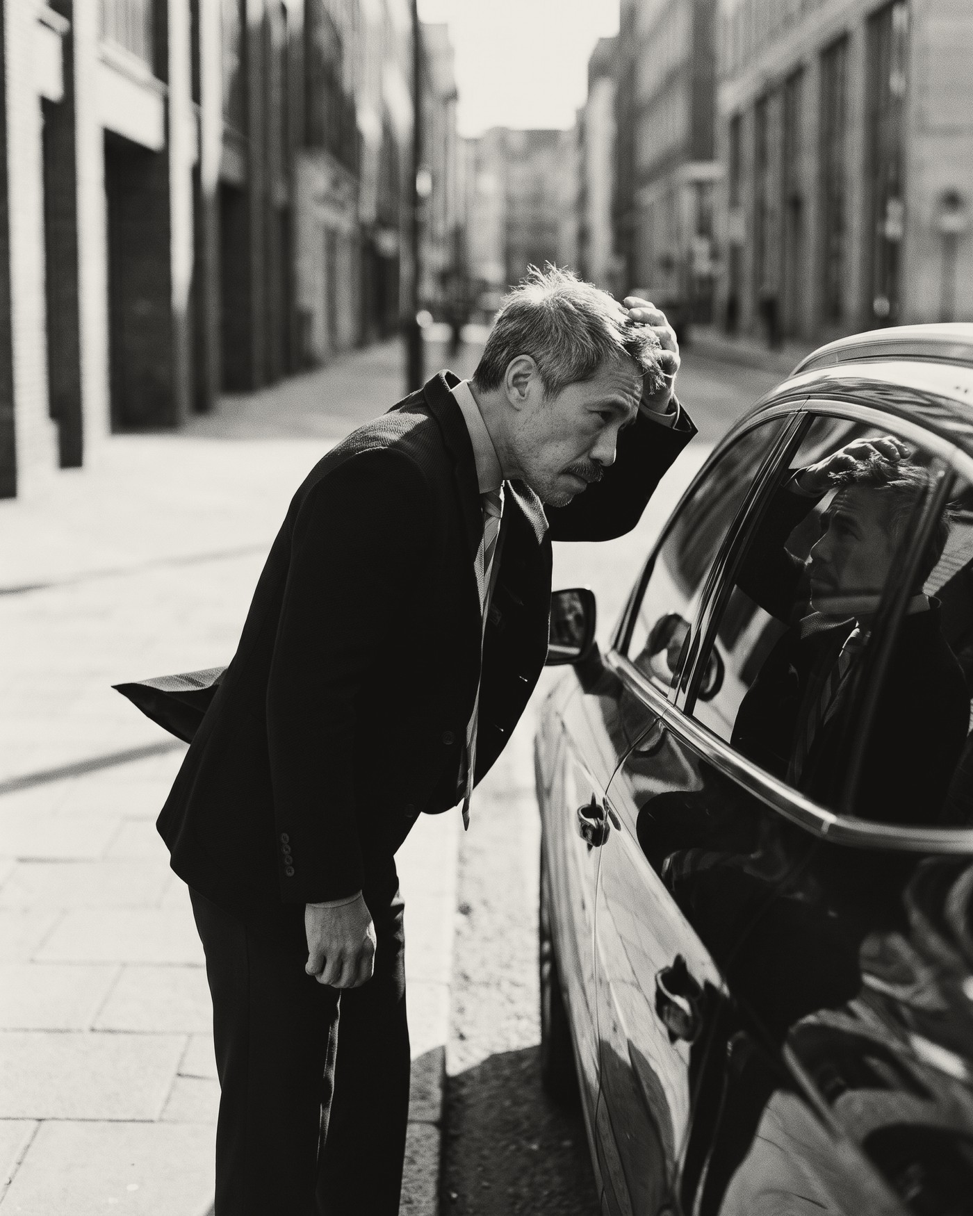 A man in a dark suit on a windy city street, bent towards the side window of a parked car, one hand pressed to the front of his hair as he checks his reflection, his jacket lifted by the wind.