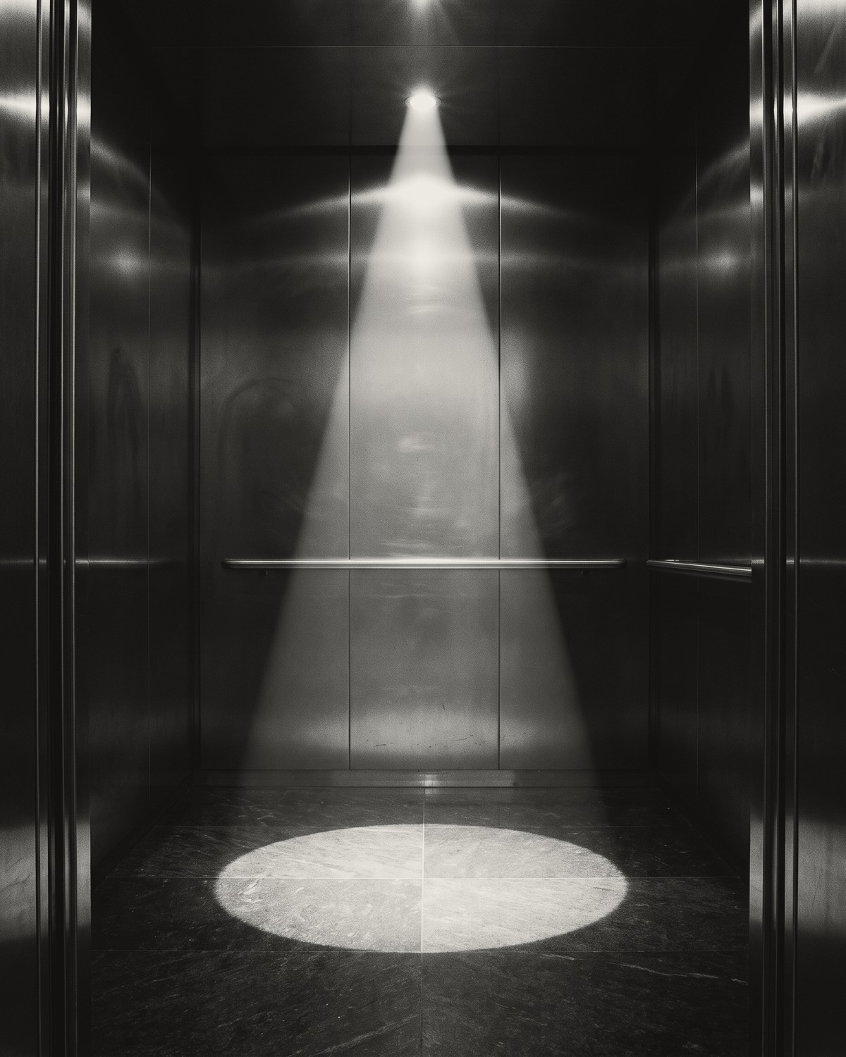 The inside of an empty lift, brushed steel walls and a handrail, a single ceiling spotlight throwing a hard cone of light onto a bright circle on the floor.