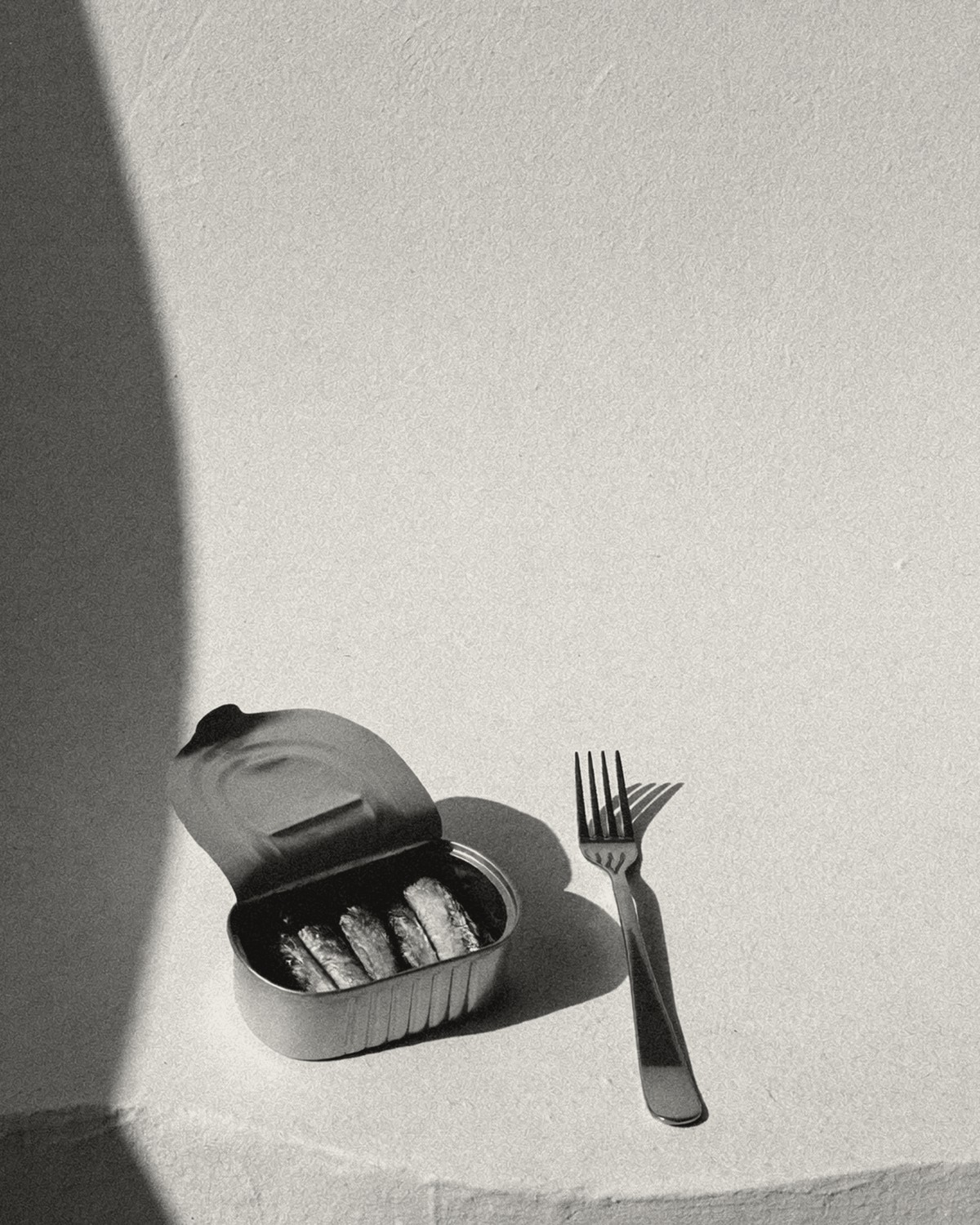 An opened tin of sardines beside a fork, on a pale table in hard sunlight.